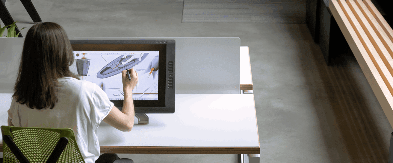 Dans un espace de travail moderne et bien éclairé, une personne aux cheveux longs est assise à un bureau et utilise un stylet pour créer une œuvre d'art numérique représentant un bateau sur un grand écran de tablette.