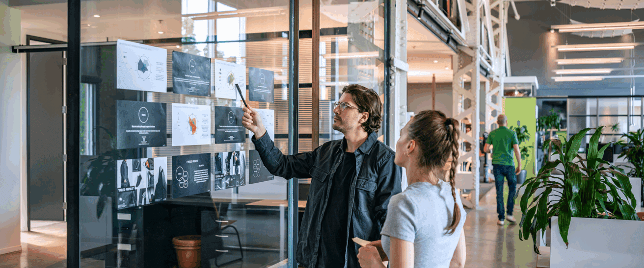 Deux personnes se trouvent dans un bureau moderne et regardent des tableaux et des documents accrochés à une paroi en verre. L'une des personnes pointe du doigt les documents tout en discutant avec l'autre. Des usines et d'autres travailleurs sont visibles à l'arrière-plan.