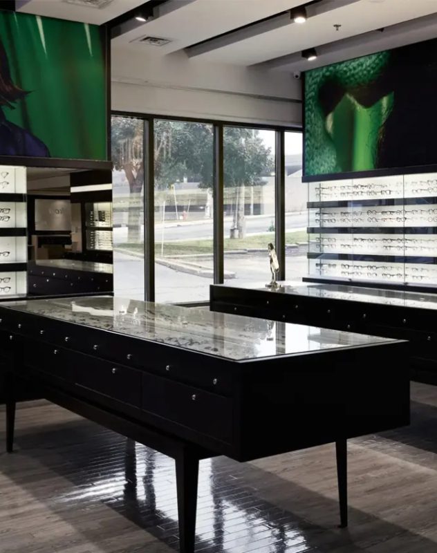 L'intérieur d'un magasin de lunettes moderne est composé de vitrines en verre contenant des montures de lunettes, de grandes fenêtres laissant entrer la lumière naturelle et d'œuvres d'art sur le thème du vert sur les murs. Le magasin semble propre et organisé.