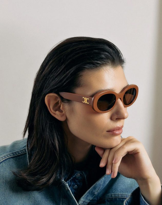 Une personne aux cheveux lisses, foncés et longs porte des lunettes de soleil ovales brunes et une veste en jean. Elle repose son menton sur sa main et regarde pensivement sur le côté, sur un fond uni.