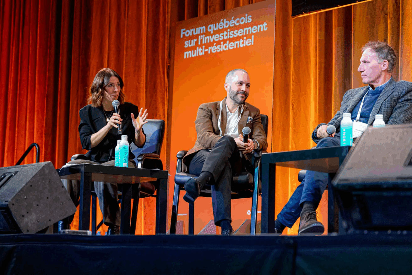 Trois personnes sont assises sur la scène du Forum québécois sur le linvestissement multi-résidentiel, avec des rideaux orange et un panneau en arrière-plan. Une femme parle dans un micro.
