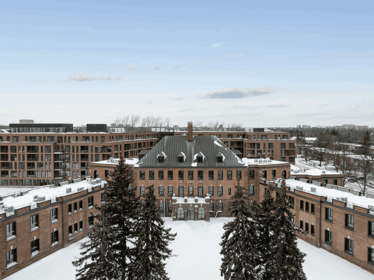 Un grand bâtiment historique en briques avec un toit vert est entouré de neige et d'arbres à feuilles persistantes, avec des immeubles d'habitation modernes à l'arrière-plan sous un ciel clair.