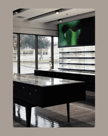 Intérieur d'un magasin de lunettes moderne avec des vitrines, des étagères garnies de lunettes, de grandes fenêtres laissant entrer la lumière naturelle et un écran numérique sur le mur affichant une image verte abstraite.