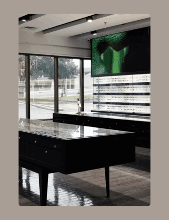 Intérieur d'un magasin de lunettes moderne avec des vitrines, des étagères garnies de lunettes, de grandes fenêtres laissant entrer la lumière naturelle et un écran numérique sur le mur affichant une image verte abstraite.