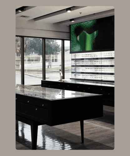 Intérieur d'un magasin de lunettes moderne avec des vitrines, des étagères garnies de lunettes, de grandes fenêtres laissant entrer la lumière naturelle et un écran numérique sur le mur affichant une image verte abstraite.