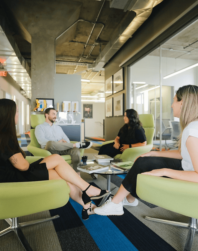 Quatre personnes sont assises sur des chaises vertes et discutent dans un bureau moderne doté de grandes fenêtres, de murs en verre et de plafonds apparents. Elles semblent détendues et sont assises autour d'une table basse.