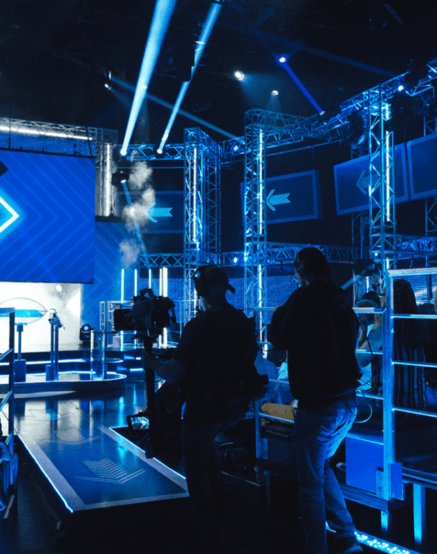Une équipe de tournage silhouettée filme sur une scène éclairée d'un bleu vibrant, avec des fermes métalliques, des écrans LED et des faisceaux d'éclairage spectaculaires, dans un studio de télévision moderne.