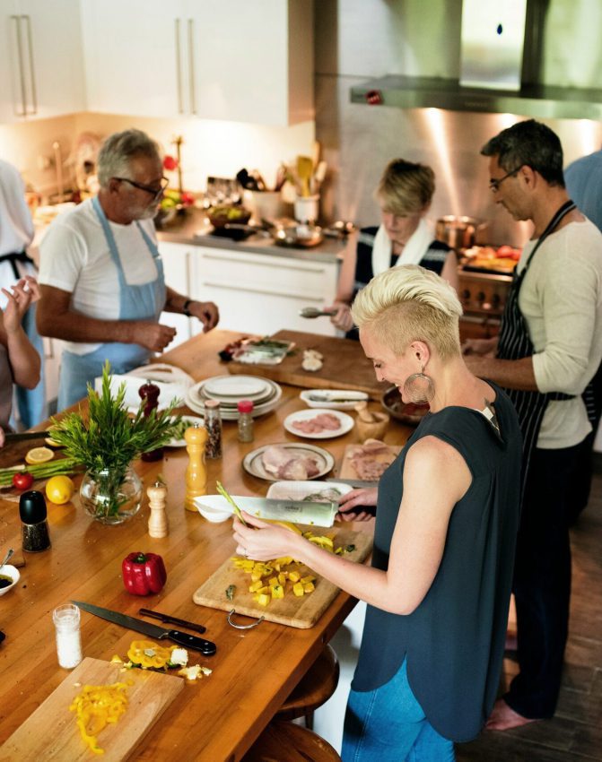 Un groupe de personnes est réuni dans une cuisine et prépare des plats ensemble. Divers ingrédients et ustensiles sont étalés sur une table en bois, et tout le monde hache et assemble des plats, créant ainsi une atmosphère animée et collaborative.