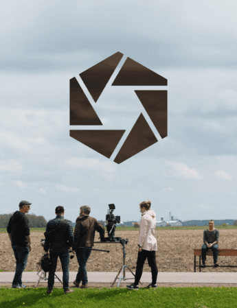 Une équipe de tournage avec des caméras et du matériel se tient devant un champ sous un ciel nuageux, tandis qu'une personne est assise seule sur un banc au loin. Un grand logo géométrique sombre est superposé au-dessus d'eux.
