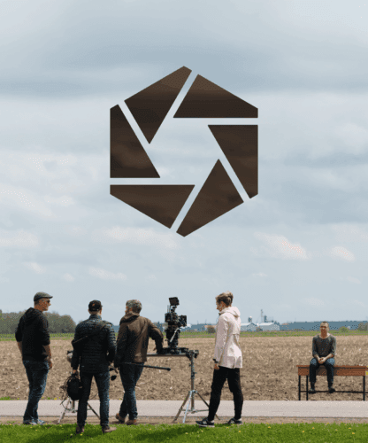 Une équipe de tournage avec des caméras et du matériel se tient devant un champ sous un ciel nuageux, tandis qu'une personne est assise seule sur un banc au loin. Un grand logo géométrique sombre est superposé au-dessus d'eux.