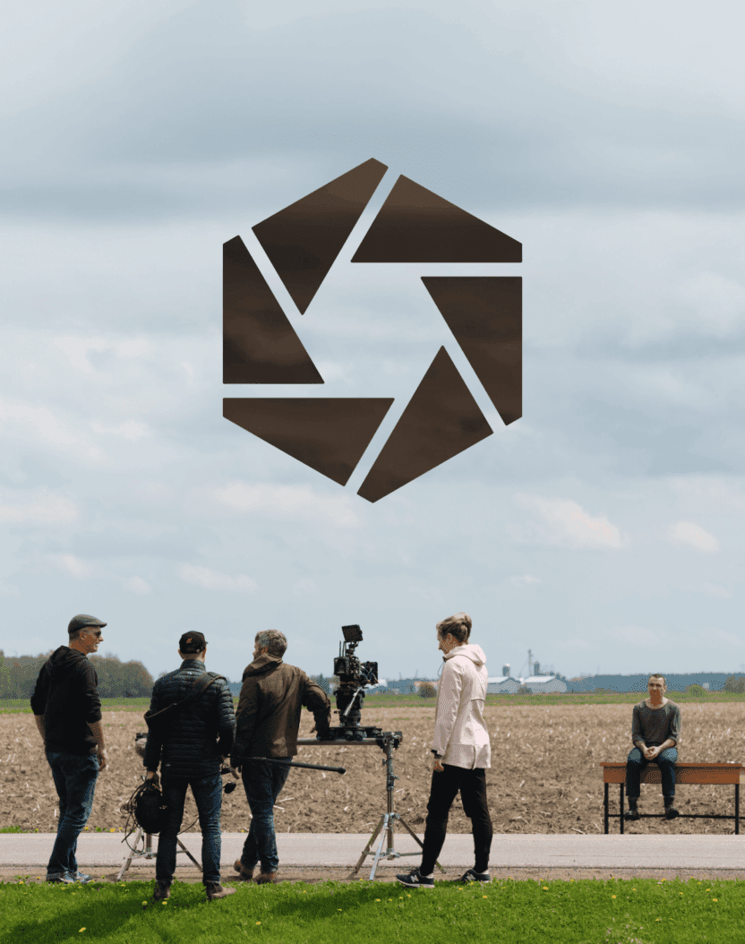 Une équipe de tournage avec des caméras et du matériel se tient devant un champ sous un ciel nuageux, tandis qu'une personne est assise seule sur un banc au loin. Un grand logo géométrique sombre est superposé au-dessus d'eux.