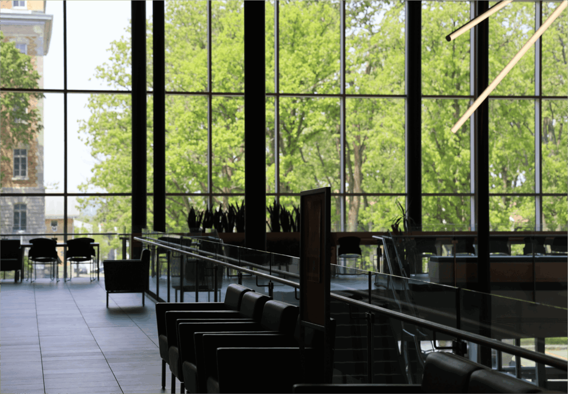 Modern indoor lounge area with black chairs and tables, large floor-to-ceiling windows revealing green trees outside, and a bright contemporary light fixture hanging from the ceiling.