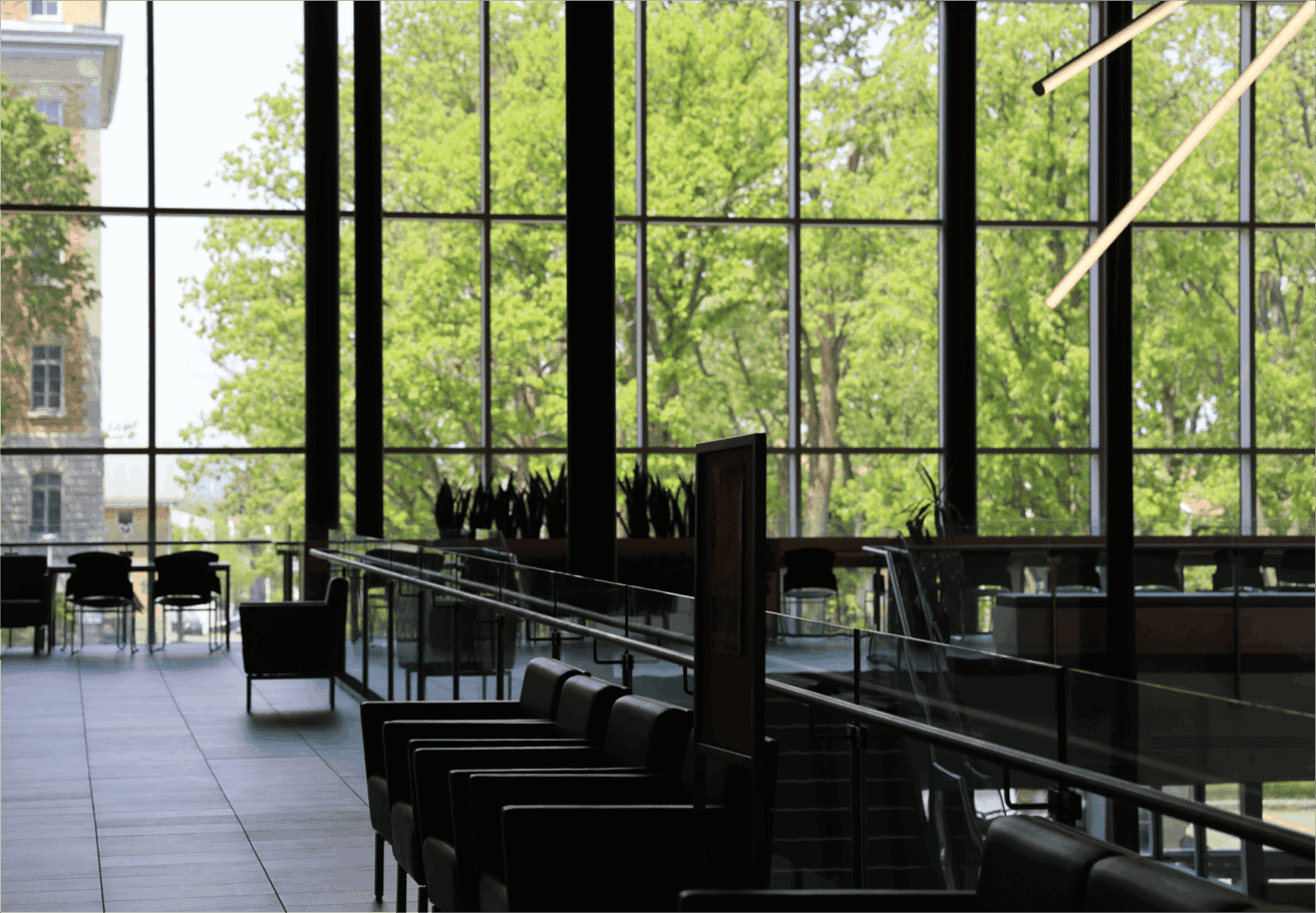 Salon intérieur moderne avec des chaises et des tables noires, de grandes fenêtres du sol au plafond révélant des arbres verdoyants à l'extérieur, et des plafonniers géométriques suspendus au premier plan.